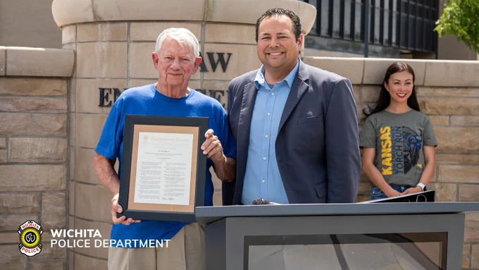 A person holding a framed certificate stands next to another person at a podium. Behind them is a smiling individual wearing a shirt with "Kansas" on it. The backdrop features a stone wall with an engraved sign that reads "Law Enforcement." The Wichita Police Department logo is visible in the foreground.