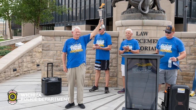 Four people stand near the Law Enforcement Memorial, wearing matching blue t-shirts. One person holds a lit torch aloft. A sign next to them reads "Torch Run." The logo for the Wichita Police Department is visible in the bottom left corner.