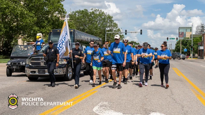 Group of people jogging down a street near a Wichita Police Department vehicle. Several participants wear blue shirts with "Torch Run" on them, and one person carries a torch. The street is lined with trees and there is a building in the background. The Wichita Police Department logo is visible in the corner.
