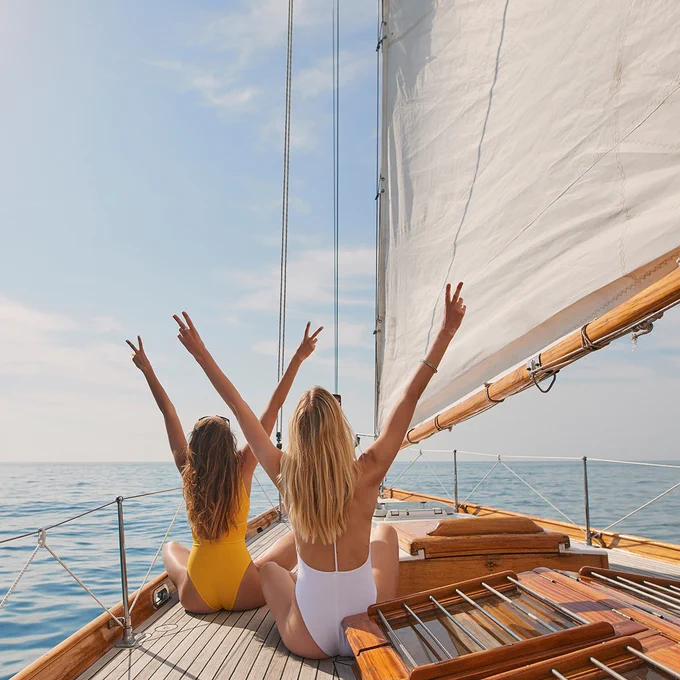 Two people sit on the deck of a sailboat, facing the ocean and raising their arms in joy. The sail is unfurled above them under a clear blue sky.