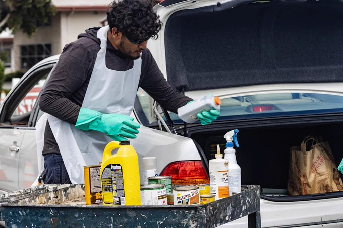 a person wearing a smock and gloves is holding a bottle of auto fluid in front of an open trunk of a car. There is a cart of various cans of paint and chemicals in front of him.