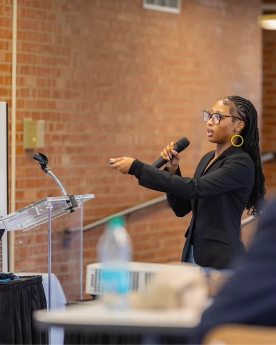 Jasmine speaking into microphone with brick wall in background