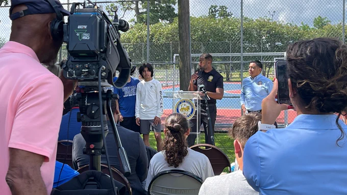 A community event is taking place outdoors at a park, where a speaker stands at a podium with the City of Houston seal, addressing an audience seated on folding chairs. A camera operator films the scene from the left side, while another person in the foreground records with a mobile device. Behind the speaker, several individuals stand near a chain‑link fence and sports court. Trees and a partly cloudy sky fill the background.
