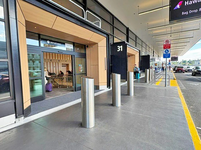 Airport terminal entrance with \"DEPARTURES\" sign and bollards, labeled \"31\", with a nearby drop-off lane.