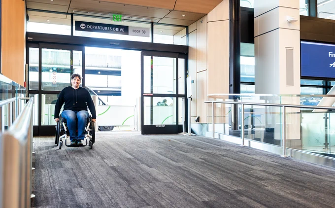 A person in a wheelchair uses a ramp in an airport terminal, heading towards automatic glass doors under a sign reading \"DEPARTURES DRIVE.\"