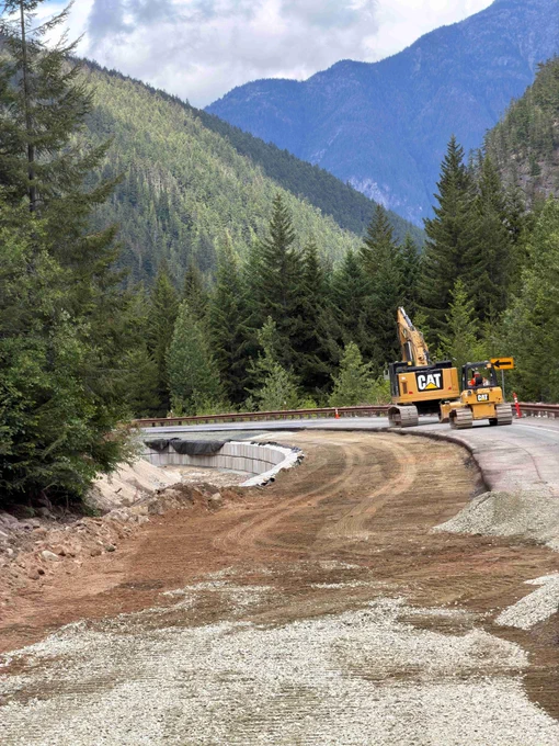 Yellow machinery repairs roadbed along a remote, mountainous section of State Route 20/North Cascades highway. A dirt section sits next to remaining pavement.