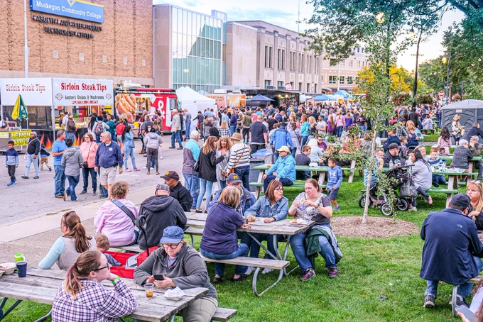 A bustling outdoor event at Taste of Muskegon features numerous food trucks and tables where people eat and socialize. The scene is lively with a variety of attendees enjoying the day, surrounded by lush greenery and buildings in the background.