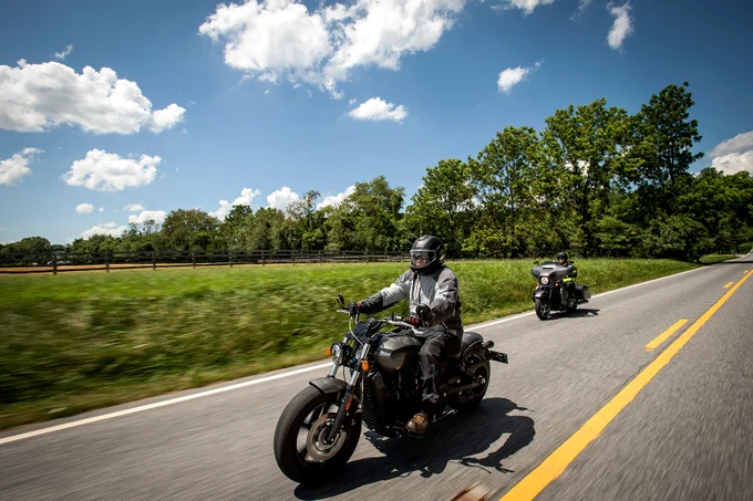 Photo of two motorcyclists wearing helmets riding down a road on a sunny day past trees and a grassy field. 