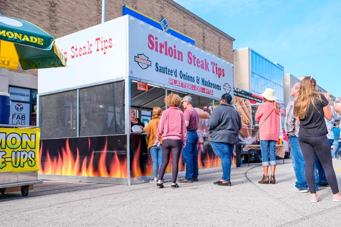 Outdoor food vendor stand titled "Sirloin Steak Tips" serving sautéed onions and mushrooms. A small group of people stands in line waiting to order. The scene is lively and takes place on a clear day.