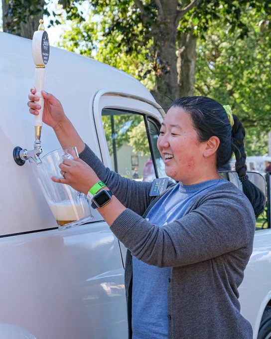 A person is pouring a beverage from a tap attached to a white vehicle at an outdoor event. They are wearing a smartwatch and standing near trees. The setting appears casual and lively.
