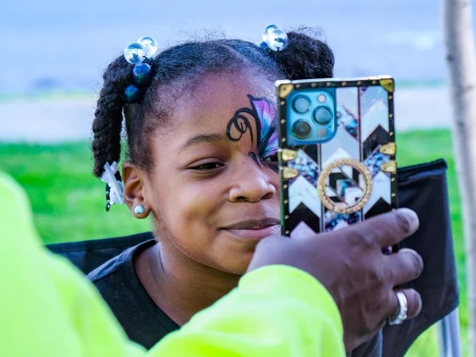 A person with butterfly face paint is smiling and looking at a smartphone with a decorative case, held by another individual. The background is outdoors with grass visible.