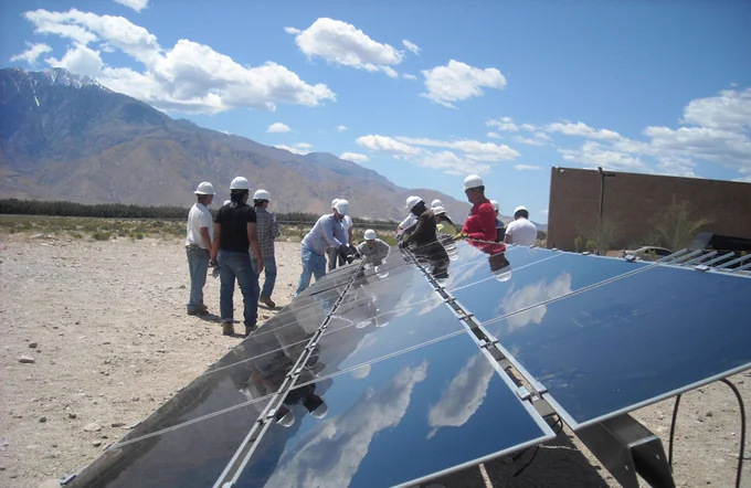 A large group of students wearing white construction hard hats stand together outdoors, closely examining a row of solar panels. The scene is set in a wide, open area with a mountainous landscape in the background. Above them, the sky is bright blue with soft, scattered clouds, creating a clean and hopeful atmosphere that highlights innovation, sustainability, and hands-on learning in renewable energy.