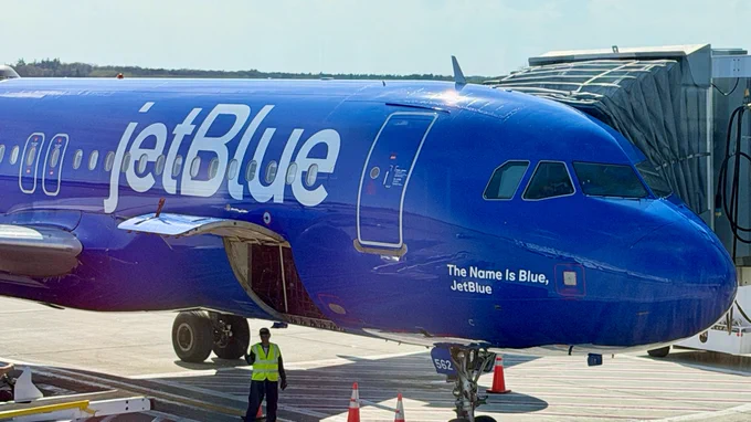 A blue-colored jetBlue airplane is parked at a gate with a jet bridge attached. The plane's unique name is "The Name is Blue, JetBlue," a nod the popular James Bond movie line.