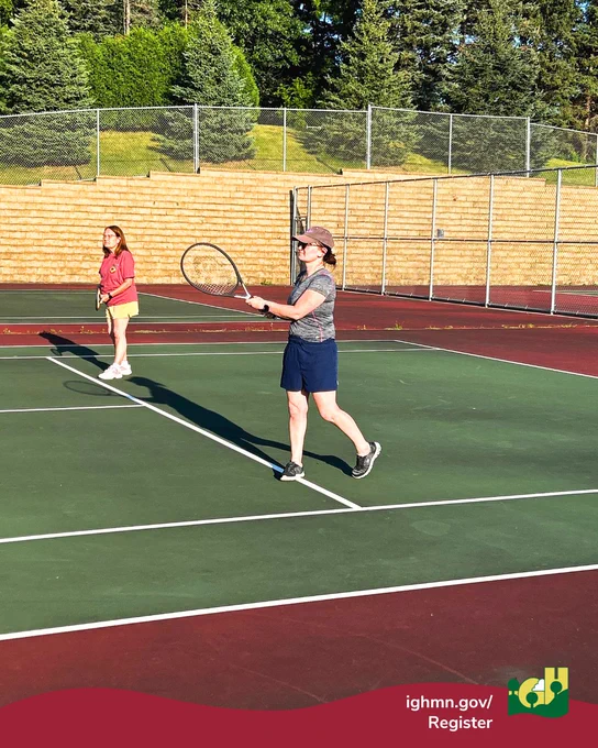 Two women playing tennis on an outdoor court surrounded by trees and a chain-link fence on a sunny day.