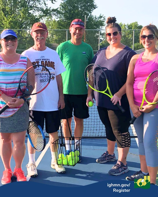 Group of five adults standing on a tennis court holding rackets with a basket of tennis balls at their feet on a sunny day.