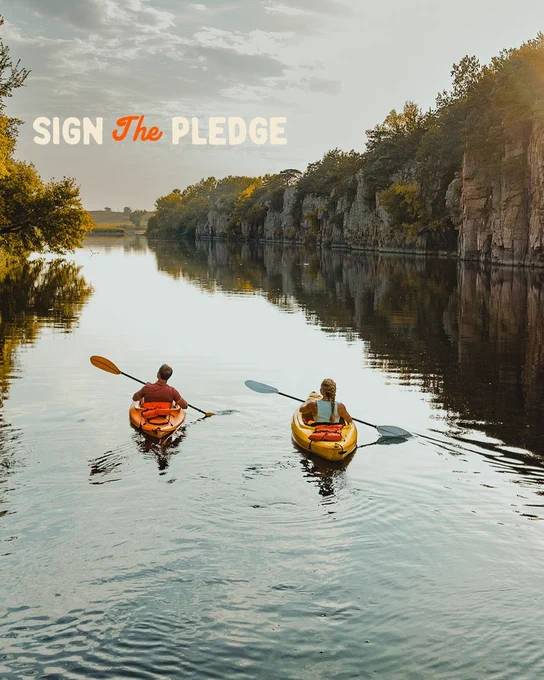 Two people kayaking on a calm river surrounded by trees and rocky cliffs, with the text "Sign The Pledge" in the sky above.