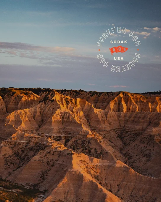 A scenic view of the Badlands National Park with rugged rock formations lit by warm sunlight. The sky is a mix of blue and clouds. A circular graphic reads "Forever 605 SoDak USA."