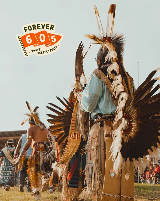 Person in traditional regalia participates in a cultural event outdoors, featuring feathered headdress and beaded adornments. A "Forever 605 Travel Respectfully" sign is visible in the background.