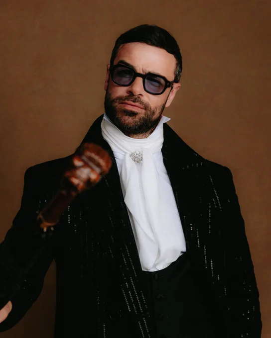 Ben Starr is photographed backstage at the BAFTA Games Awards with Google Play 2026. He wears a black sequinned suit with a white shirt and ruffled scarf. He holds an umbrella up, and pulls a satisfied smirk.