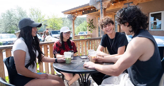 Four people sit at an outdoor café table, holding drinks and smiling. A building with a wooden sign that says "Cabra Coffee" is in the background. Cars and greenery are also visible.