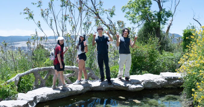 A group of four people standing on a stone path near in Carlito Springs, surrounded by lush greenery and distant hills in the background. They are smiling and making Lobo hand signs. The scene is bright and sunny.