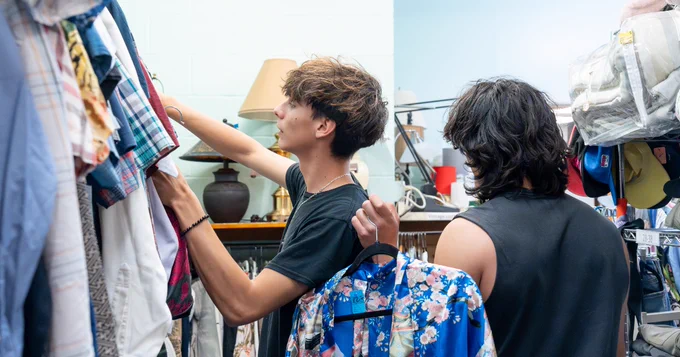 Two people are shopping in Talking Talons thrift store, browsing through racks of various shirts. One is holding a colorful floral shirt on a hanger. The store is filled with a variety of garments and accessories, and a lamp and hats are visible in the background.