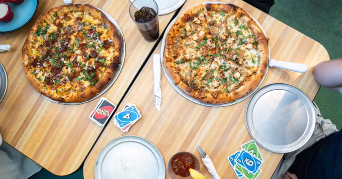 At Trail Rider Pizza, there are two pizzas on a wooden table, accompanied by drinks. A game of UNO is in progress, with cards and trays nearby.