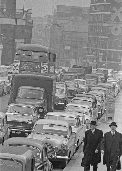 A black and white photo of a busy city street with a traffic jam. Numerous cars are lined up, and a double-decker bus is visible in the middle. Two individuals in suits and hats are walking along the sidewalk. The surrounding buildings exhibit classic mid-20th century architecture.