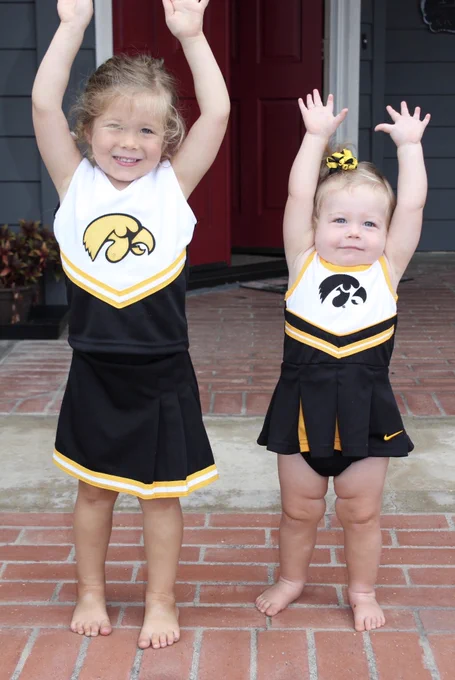 Two young children standing barefoot on a brick porch, dressed in matching University of Iowa cheerleading outfits with raised arms, in front of a red door and gray siding.

Photo by: Karen Potter