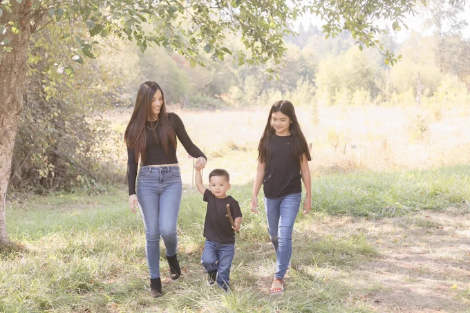A woman, a young girl, and a toddler boy walk together in a sunny, grassy field. The woman, smiling, holds the boy's hand as he carries a stick. All three have dark hair and wear black tops with blue jeans. Green trees and bushes blur in the background.