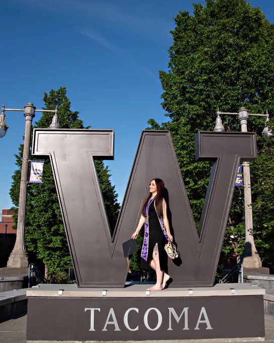 Smiling woman in a black dress and purple and gold graduation stole stands barefoot on a large dark grey 'W' structure. She holds a graduation cap and shoes. Below the 'W' is 'TACOMA' in silver letters. Bright blue sky and green trees are in the background.