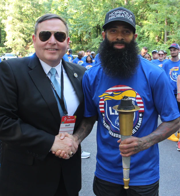 Man in suit shaking hands with a bearded man wearing a Norfolk Probation cap holding a ceremonial torch outdoors.