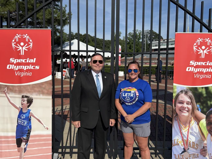 Two people stand and smile in front of an iron gate with Special Olympics Virginia signs on either side of them. 