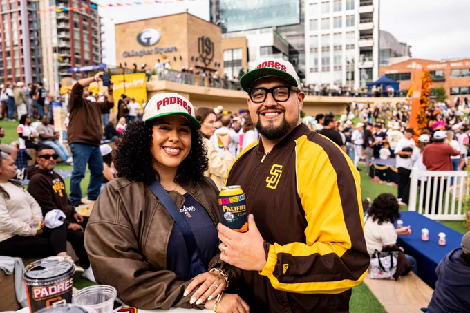 Photo of two Padres fans in Gallagher Square during Party in the Park. They are both wearing Padres hats that are white, red, and green for Mexican Heritage Night. They are also holding up a Pacifico beer.