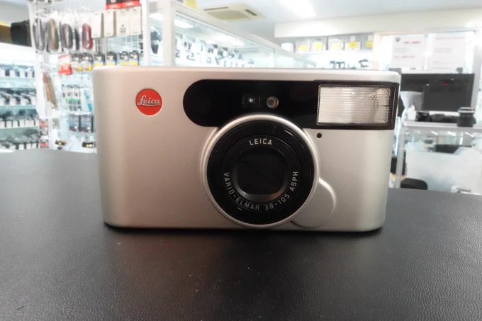 ​レモン社銀座店に入荷したコンパクトフィルムカメラ「ライカ C1（Leica C1）」の外観写真。スタイリッシュなシルバーのアルミニウム製ボディで、正面にはライカを象徴する赤いロゴ（赤バッジ）が配置されています。1999年発売の、近未来感のある洗練されたインダストリアルデザインが特徴的なカメラです。
​
​An exterior photo of the Leica C1, a compact film camera newly in stock at Lemon-sha Ginza store. It features a stylish silver aluminum body with the iconic red Leica badge on the front. This camera is known for its futuristic and sophisticated industrial design, originally released in 1999.