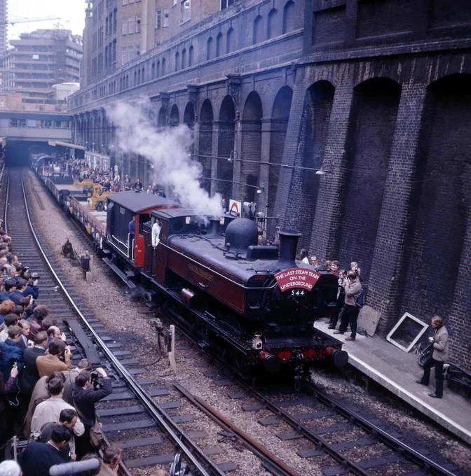 An ex-GWR pannier tank 0-6-0 steam loco hauls a brake van and a selection of engineers wagons westbound through Barbican on the unelectirifed 'Widened Lines' with a large number of people on both platforms taking photograps.