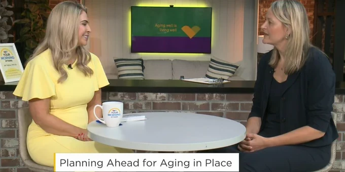 TV interview of two women seated at a round table discussing aging in place with sign and a screen behind them.