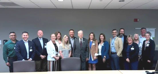 A group of people standing together in a conference room, smiling. There is a mix of formal and business casual attire. They are posing in front of a table with a neutral-colored wall in the background.