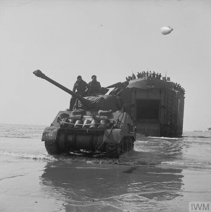 A tank exits a British landing craft onto a beach during a military operation. The landing craft has the number "5506" visible. A barrage balloon floats in the sky above.