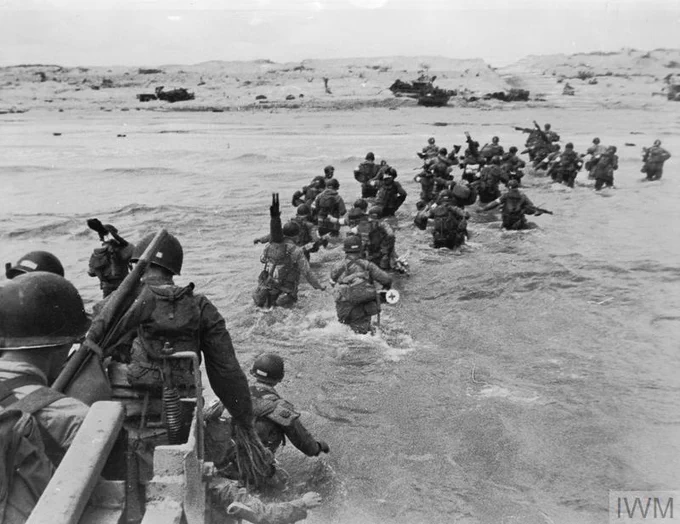 A group of soldiers wade through the shallow water towards the shore during the D-Day landings at Normandy in World War II. The beach is visible in the background, along with amphibious vehicles. The soldiers are equipped with helmets and gear as they advance.