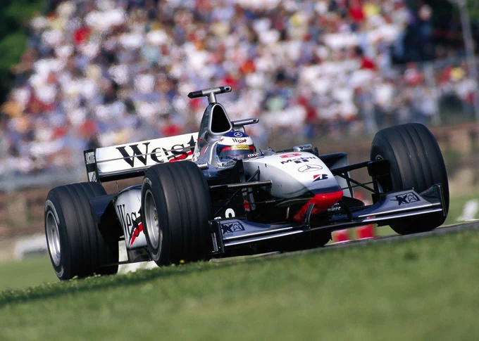 A Dutch angle shot canted slightly to the left of Mika Häkkinen on track in his grey, black and red, West sponsored McLaren MP4-13. The front and rear wheels are much more similar width, with a higher nose, air intake and lower rear wing. 1998 San Marino Grand Prix.
