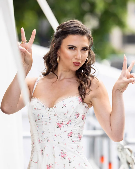 A woman standing backstage at West End LIVE in a floral dress pouting and holding up two peace signs.
