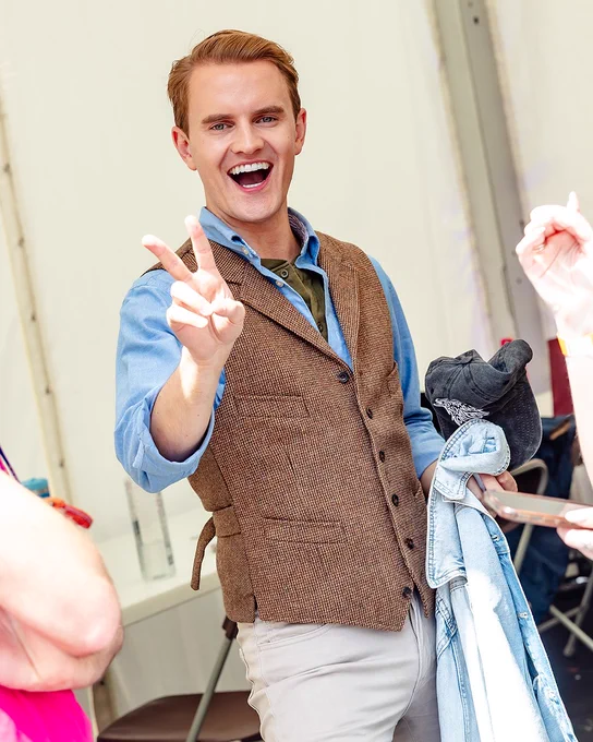 A man standing backstage at West End LIVE in a waistcoat and shirt smiling and holding up a peace sign. 