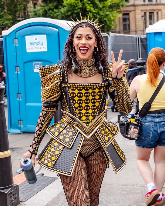 A member of Six The Musical walking backstage at West End LIVE a Tudor-inspired costume smiling and holding up a peace sign.