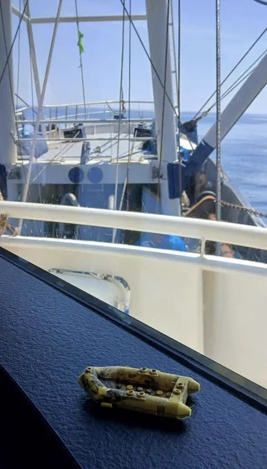 Picture shows a yellow Lego life raft on a black textured surface. The picture has been taken on board a fishing vessel, which is visible in the background.