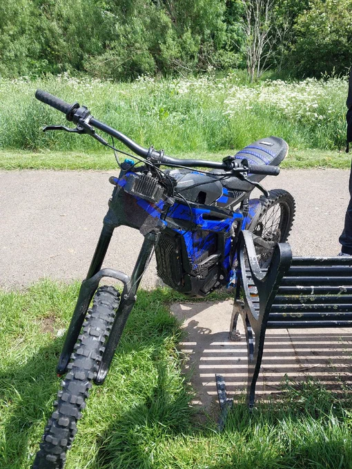 A blue and black electric dirt bike parked on a path next to a park bench with a grassy background.