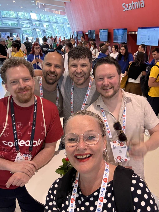 A group of five smiling people posing for a selfie at a crowded indoor event, with a large red wall in the background that has the word "Szatnia" written on it.