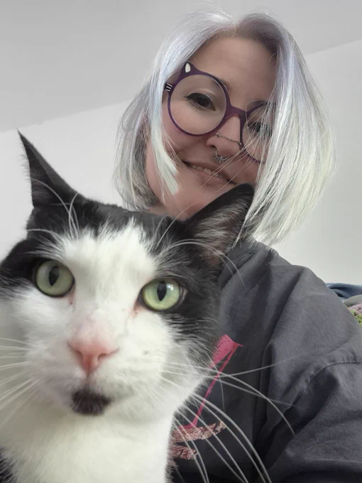 Dougie, the black and white three-legged none tail cat, is looking at the camera, and his mummy is behind him. She has short white-ish hair and nose rings and cat glasses.