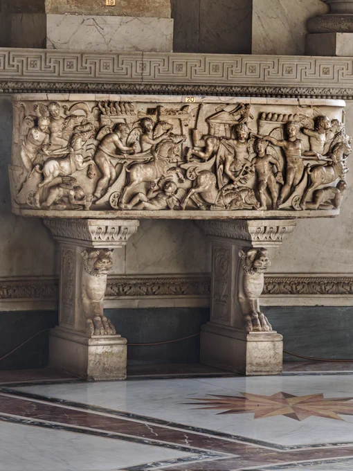 SARCOPHAGUS FRONT WITH CHARIOT RACE, 160-170 CE. VATICAN MUSEUMS

The high-relief scene depicts winged Erotes driving four-horse chariots in a chaotic race around the Circus Maximus spina, complete with miniature turning posts (metæ) and a dramatic chariot crash in the centre. In a Roman child's funerary context, the race symbolizes the course of human life, and the crash represents an untimely death. Modified in the 1780s by restorer Antonio Pazzaglia to fit Pope Pius VI’s neoclassical museum design, the original rectangular box was sawn into a flat slab, and its side edges were beveled into a curved profile. All original Roman paint was scraped off to achieve a pristine white marble look. The panel is mounted as a console table on two unrelated ancient marble table legs or trapezophora, which feature high-relief, roaring lion heads tapering down into muscular legs and clawed paws resting on flat bases.