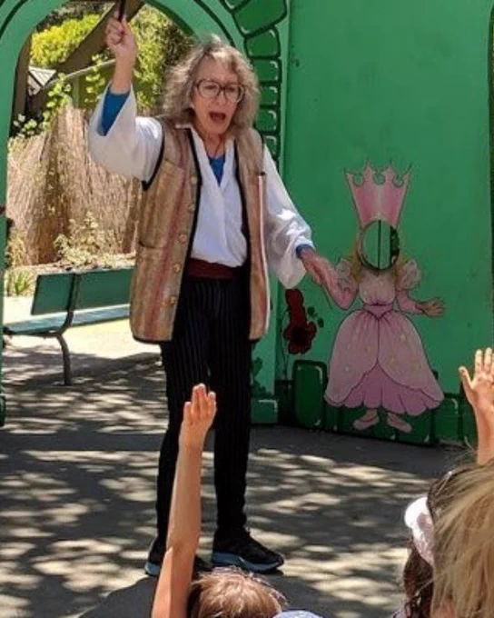 Magician stands on an outdoor staged, with wand raised. People in front of her are raising their hands.